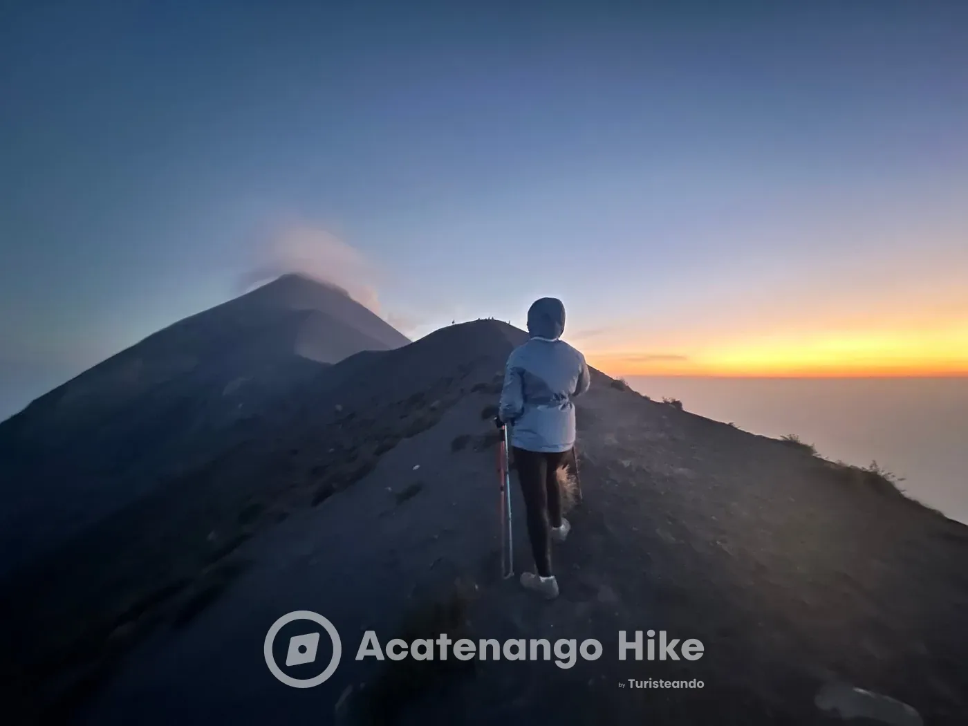 Hiker on the Acatenango summit ridge at dusk with Volcán de Fuego ahead